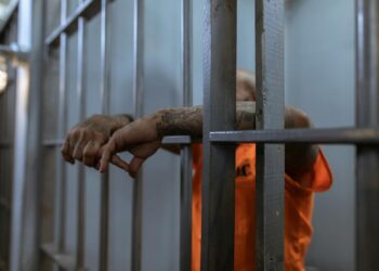 A tattooed inmate's arms rest on jail cell bars, wearing an orange uniform inside a prison.