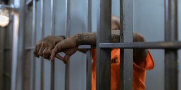 A tattooed inmate's arms rest on jail cell bars, wearing an orange uniform inside a prison.