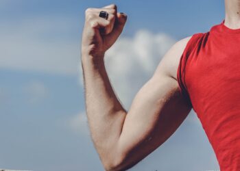 Close-up of a flexed arm in a red shirt against a clear blue sky, symbolizing strength and empowerment.