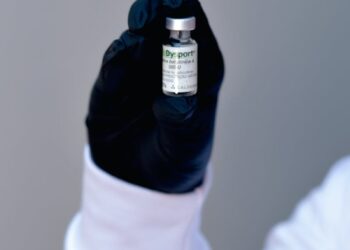 A healthcare professional's gloved hand holding a small medicine vial with a blurred background.