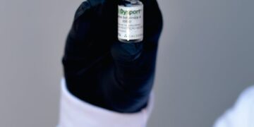 A healthcare professional's gloved hand holding a small medicine vial with a blurred background.