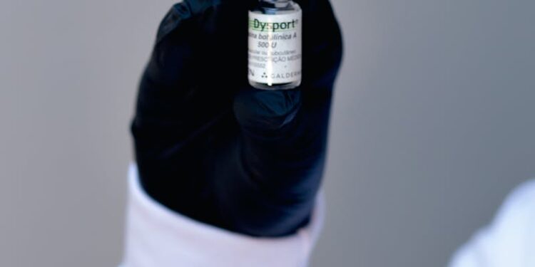 A healthcare professional's gloved hand holding a small medicine vial with a blurred background.