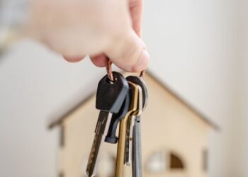 Close-up of a hand holding keys with a miniature wooden house in the background, symbolizing real estate investment.