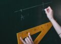 Two hands drawing a line with chalk on a blackboard using a triangle ruler